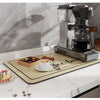 A silver coffee grinder and a natural fiber coffee mat are displayed on a white countertop.