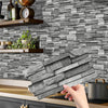 A hand holds a stack of gray stone tile stickers against a gray stacked stone wall background.