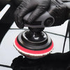 A hand holds a black polishing pad with a red and white wool surface above a reflective black surface.