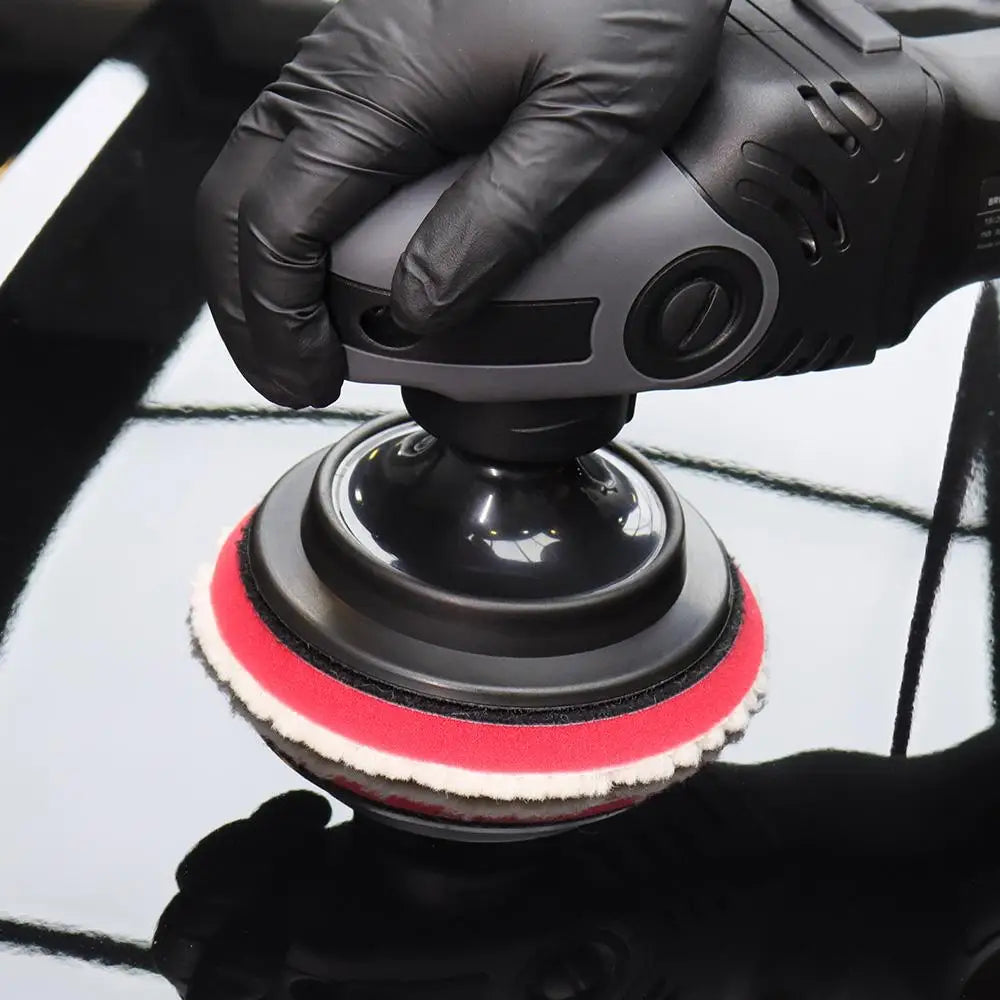 A hand holds a black polishing pad with a red and white wool surface above a reflective black surface.