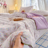 A hand is holding a light purple floral quilt blanket on a bed with soft lighting.