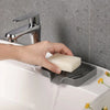 A hand is using a silver faucet to dispense water into a self-draining silicone soap tray on a white sink.
