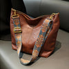A brown leather shoulder bag with golden embroidery and patterned leather straps displayed on a gray surface.