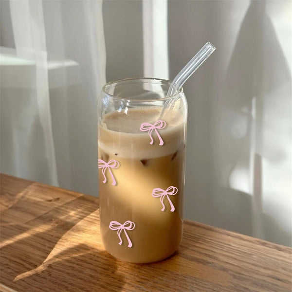 كوب زجاجي مملوء بالقهوة المثلجة أو الشاي، مزين بشرائط فراشة وردية، موضوع على سطح خشبي.