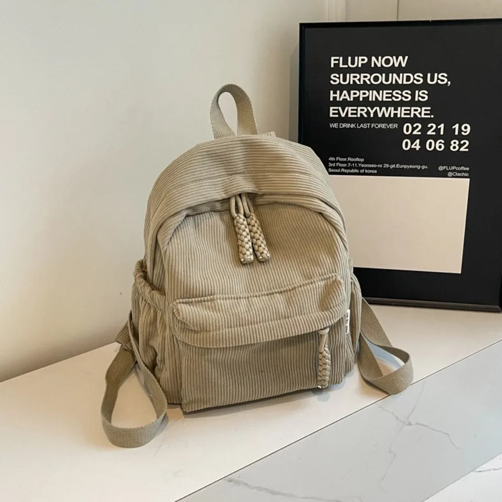 A beige corduroy casual backpack is displayed on a white surface next to a framed advertisement.