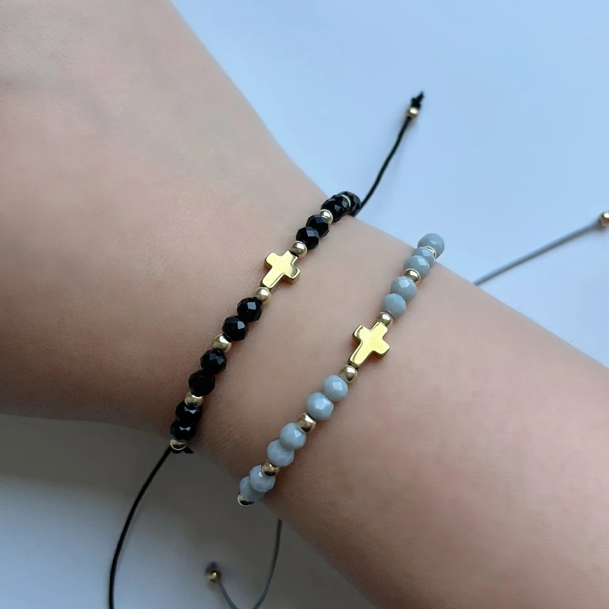 Bracelet perlé noir et bleu clair avec croix dorée porté au poignet.
