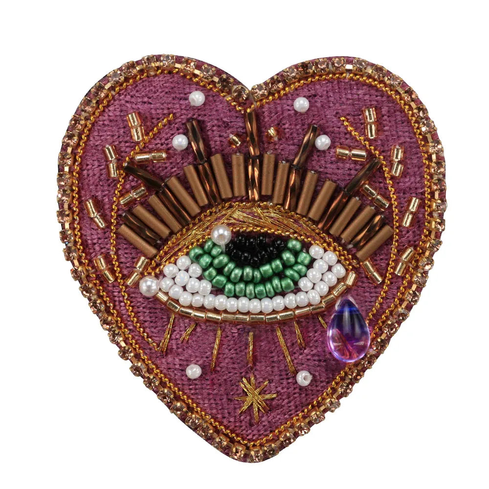 A heart-shaped patch featuring a detailed eye design made of gold and white beads on a purple fabric background.