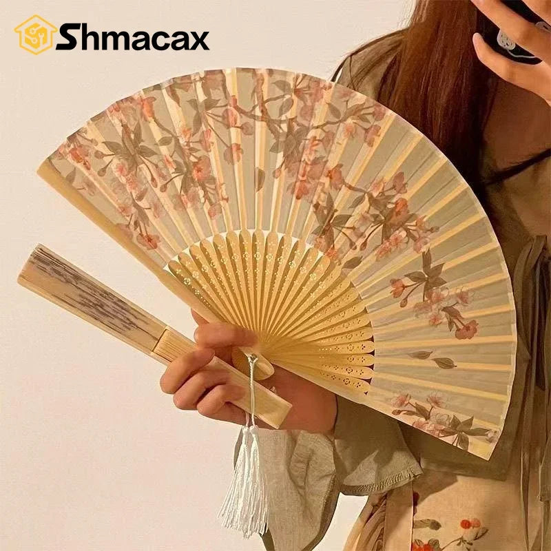 A hand holds an open bamboo and silk folding fan with a floral design against a light background.