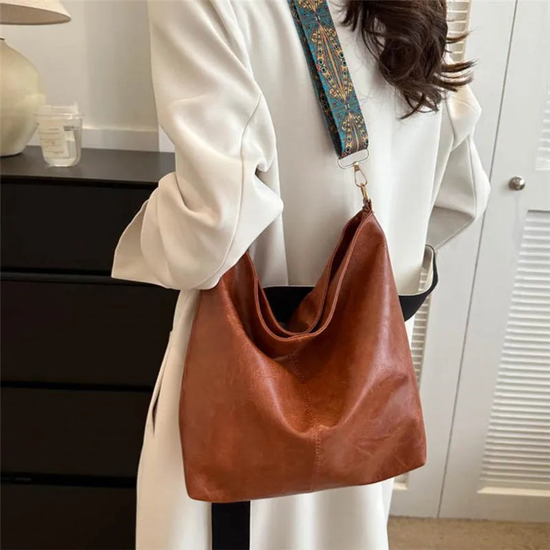 A woman holds a brown leather shoulder bag with gold embroidery and a patterned strap against a white coat.