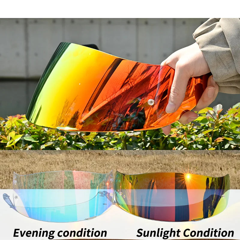 A hand holds an orange motorcycle helmet visor lens shield showing a vibrant sunset reflection.