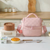 A pink insulated lunch bag is displayed on a table next to a food container and cutlery.