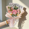 A hand holds a bouquet of pink and yellow handmade crochet artificial flowers wrapped in sheer pink paper.