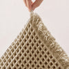 Close-up of a beige waffle weave bath mat with fluffy tassels being held by a hand.