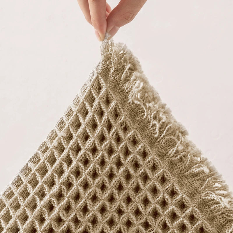 Close-up of a beige waffle weave bath mat with fluffy tassels being held by a hand.