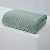 A folded, textured, sage green bath towel with a chevron pattern resting on a white surface.