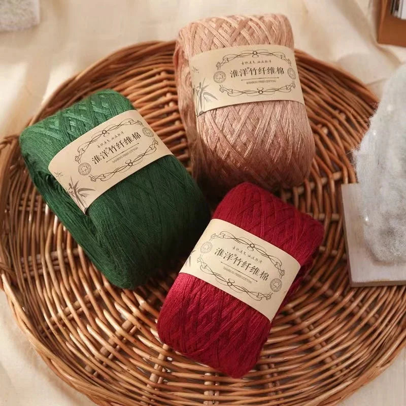 Three skeins of pure cotton yarn in shades of green, beige, and red are displayed on a woven tray.