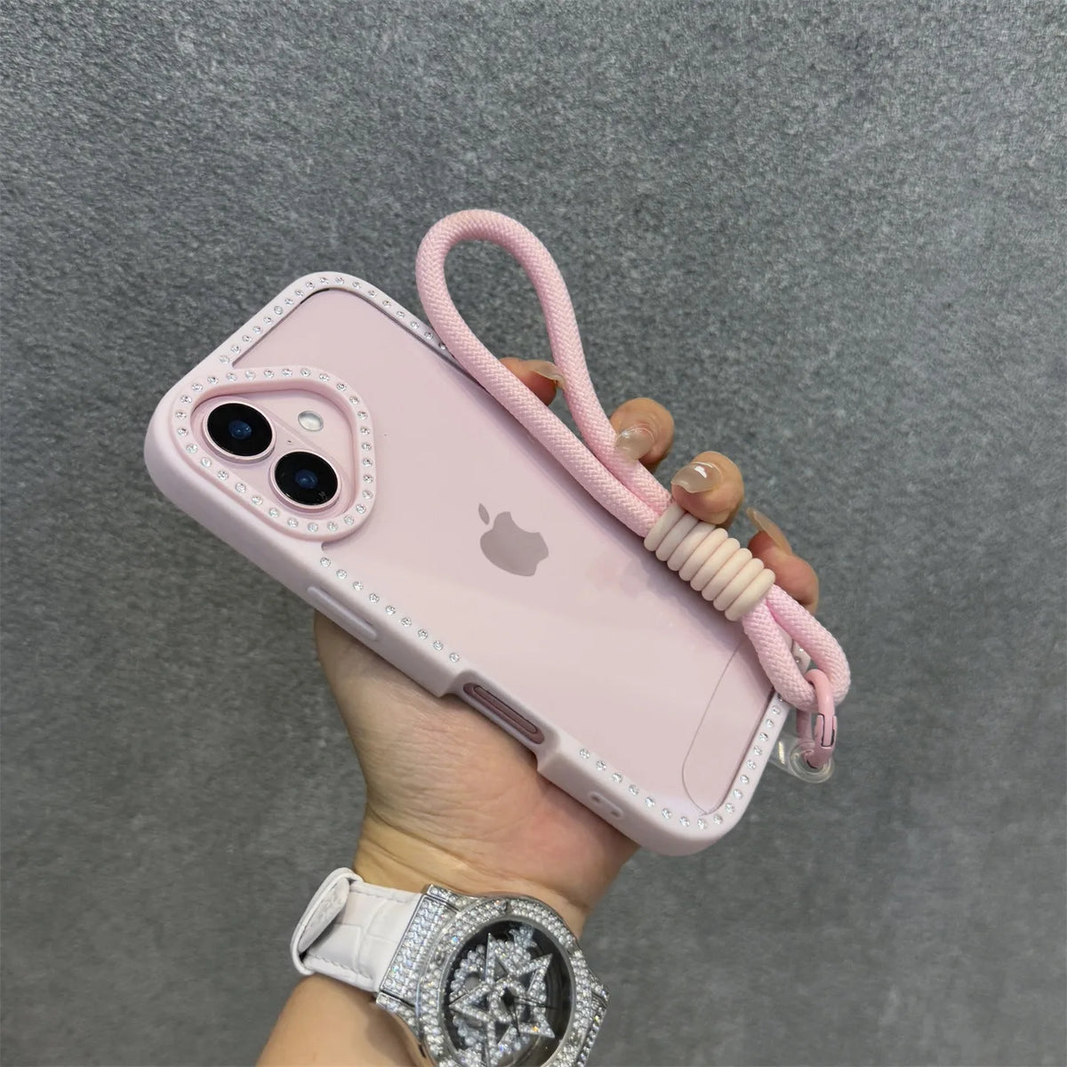 Una mano sostiene una funda de teléfono rosa con pulsera y un reloj de diamantes plateado contra un fondo gris.