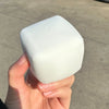 A hand holds a white, square squishy milk tofu stress relief fidget toy against a gray outdoor background.