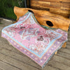 A pink and purple bohemian jacquard woven throw blanket draped over a wooden log on a deck.