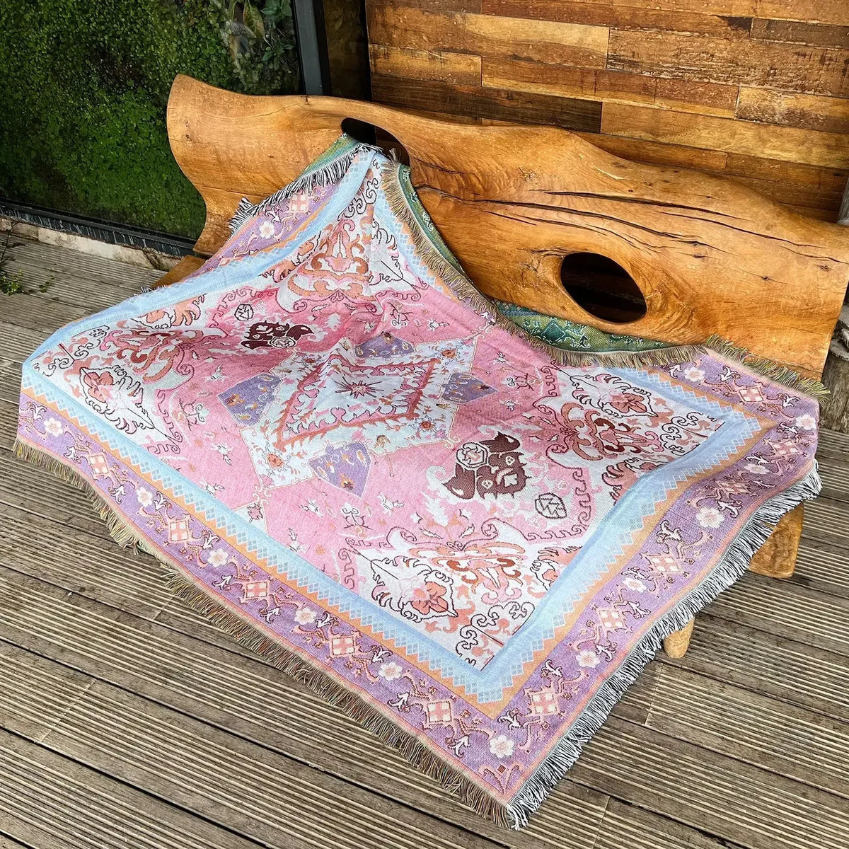 A pink and purple bohemian jacquard woven throw blanket draped over a wooden log on a deck.