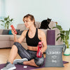 A woman sits on the floor with a black laundry bag and pink phone in a bright living room.