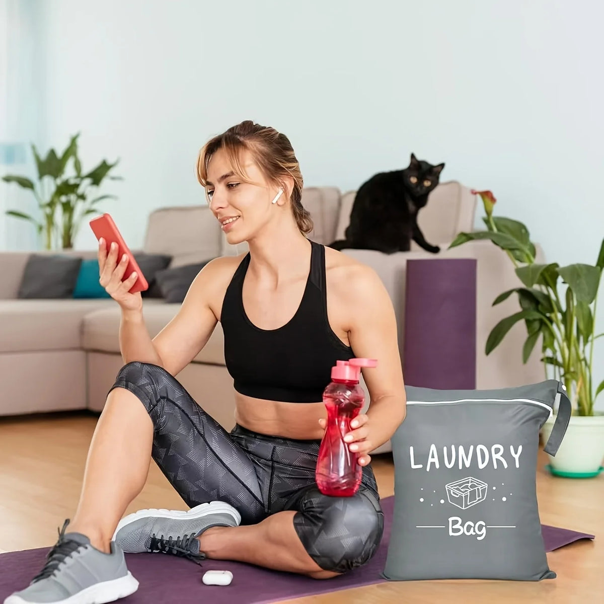 A woman sits on the floor with a black laundry bag and pink phone in a bright living room.