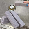 Several gray foam glass pumice stones are displayed on a metal surface next to a glass cleaning brush.