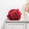 A deep red, hand-knotted velvet throw pillow resting on a white wooden shelf next to a light-colored decorative object.