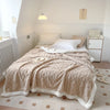 A beige fleece throw blanket is draped over a white bed with light-colored bedding and a wooden nightstand.
