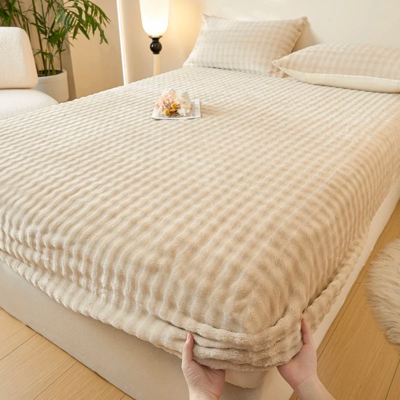 A beige faux fur fitted sheet is being held, displayed on a bed with white linens and natural light.
