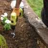 A person is using a yellow garden auger drill bit in dark soil next to a concrete edge.