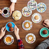 A flat lay of various coffee cups, cookies, and a sparkling letter-themed diamond painting coaster set on a wooden table.