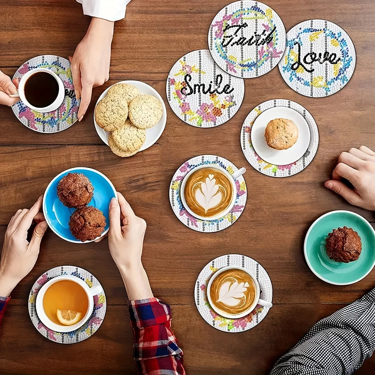 A flat lay of various coffee cups, cookies, and a sparkling letter-themed diamond painting coaster set on a wooden table.