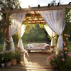 A wooden pergola draped with sheer white curtains frames a garden area featuring lush greenery and flowering plants.