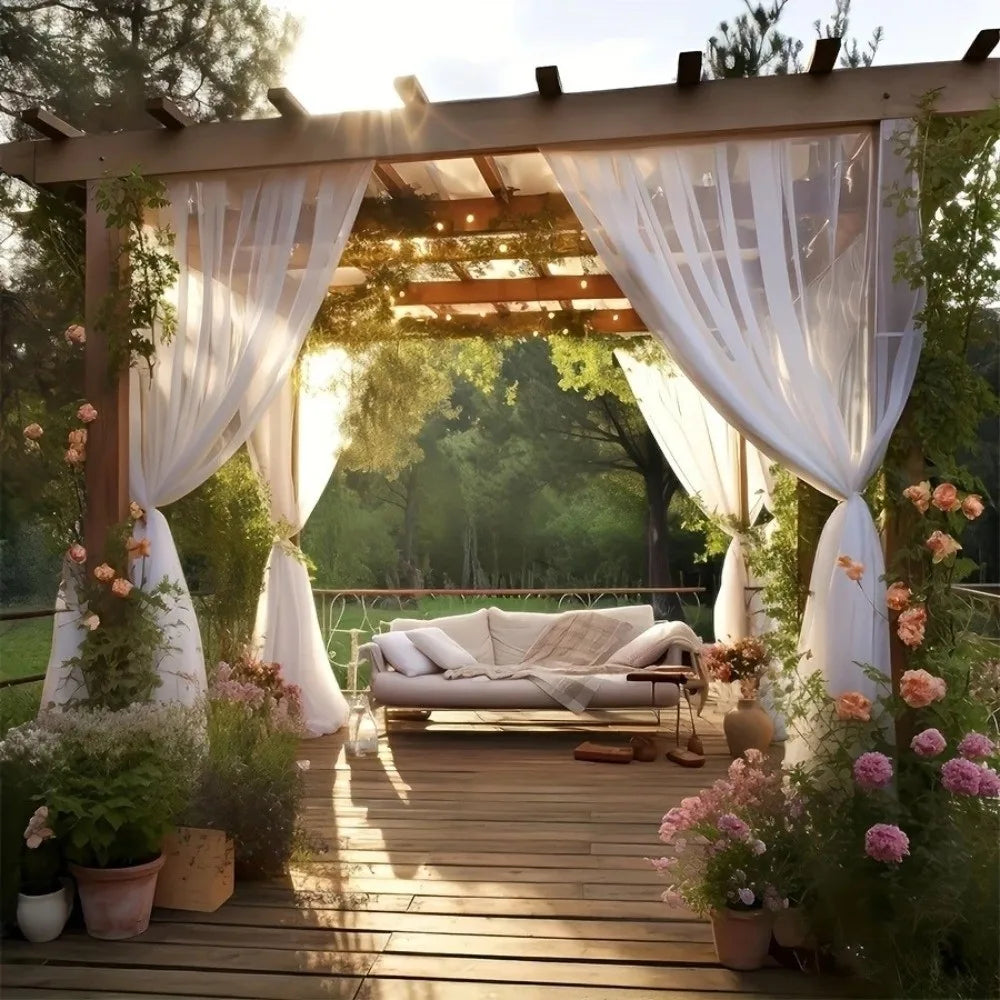 A wooden pergola draped with sheer white curtains frames a garden area featuring lush greenery and flowering plants.