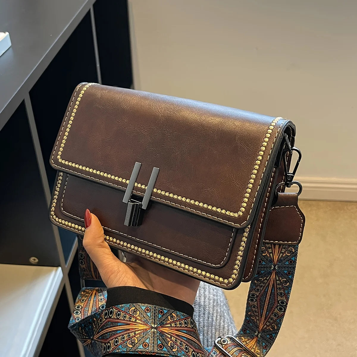 A brown leather mini cross-body bag with gold stud detailing and a patterned strap is being held by a hand.