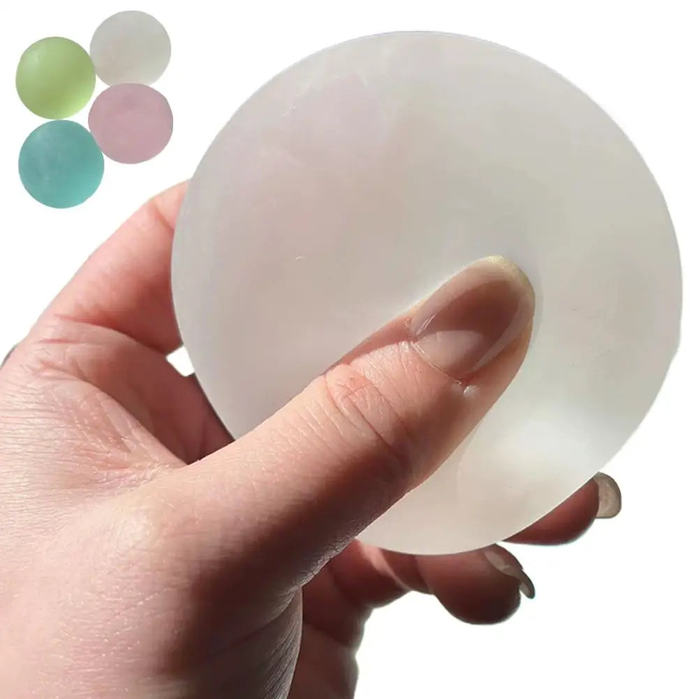 A hand is squeezing a translucent white coconut oil slow rising squeeze ball, with several colored oil balls visible nearby.