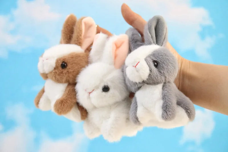 A hand is holding three soft plush bunny toys: brown, white, and gray.