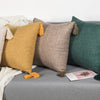 A collection of linen throw pillows in mustard yellow, beige, and deep green with tasseled edges on a gray surface.