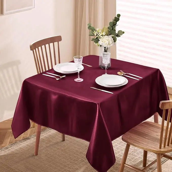 A deep burgundy satin tablecloth is set for dining with white plates and silverware, featuring a floral centerpiece.