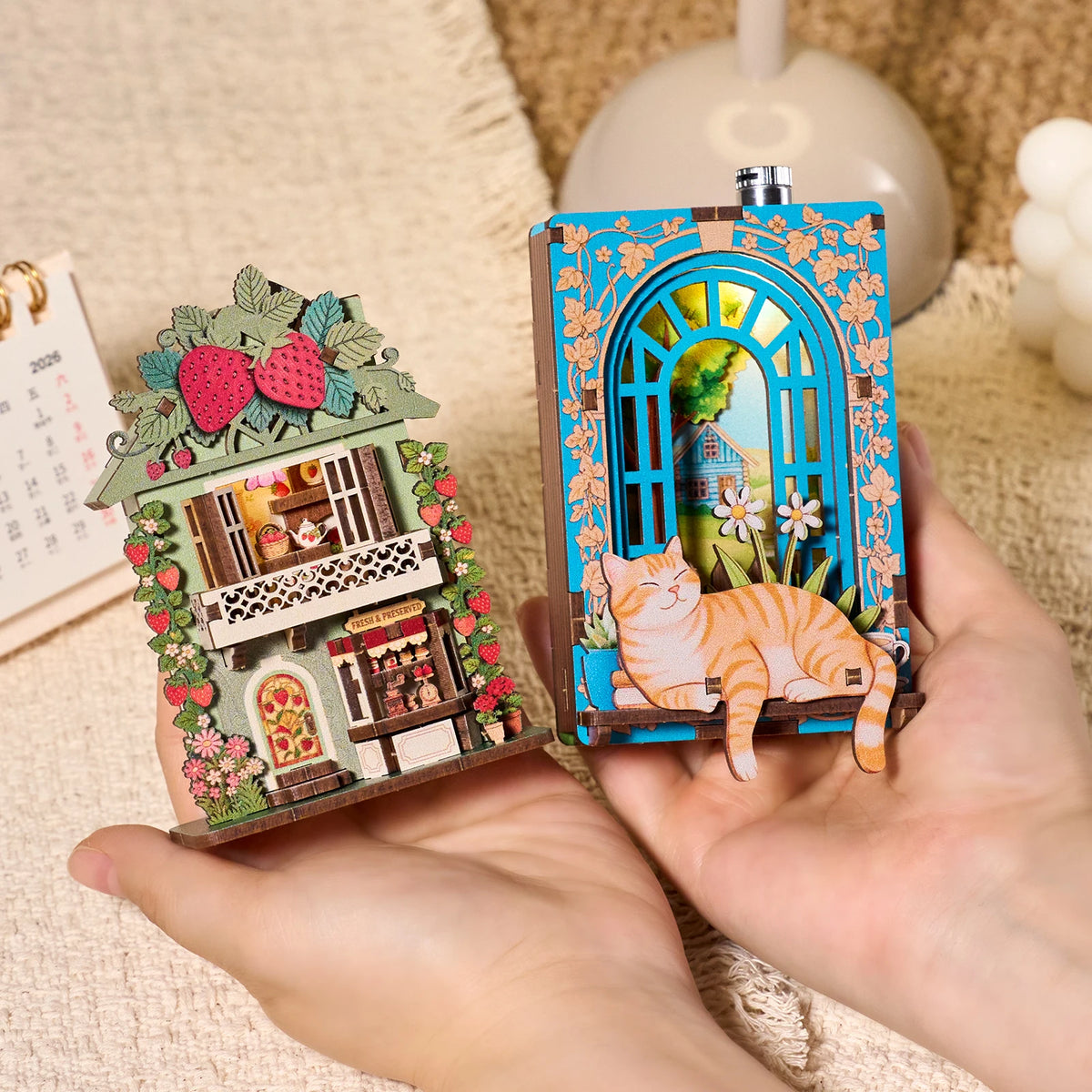A hand holds a DIY book nook fridge magnet kit featuring a wooden house with floral accents and an arched window display.