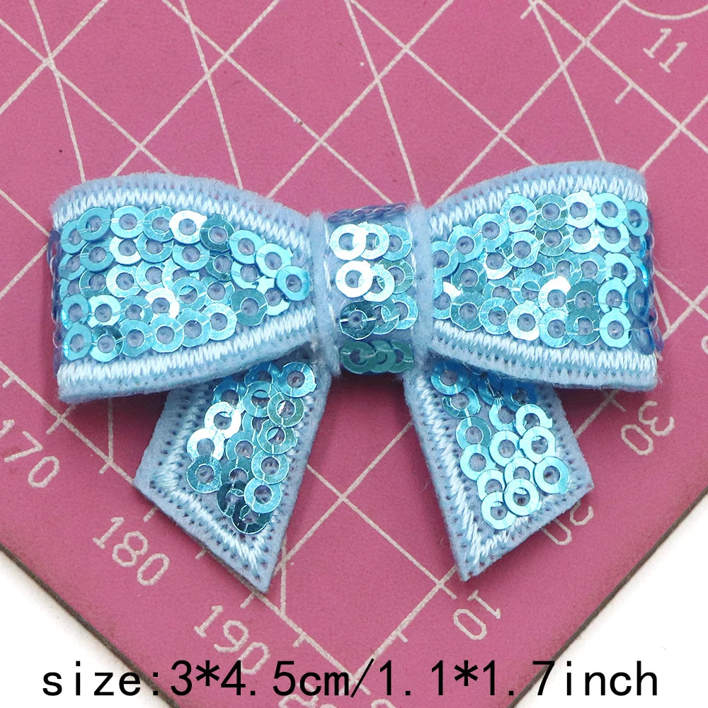 A light blue sequin bow made of fabric is shown on a pink background.