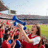 Eine junge Frau in einem roten Sporttrikot bläst eine blaue portable Luftpumpe-Sicherheitshupe während eines Fußballspiels.