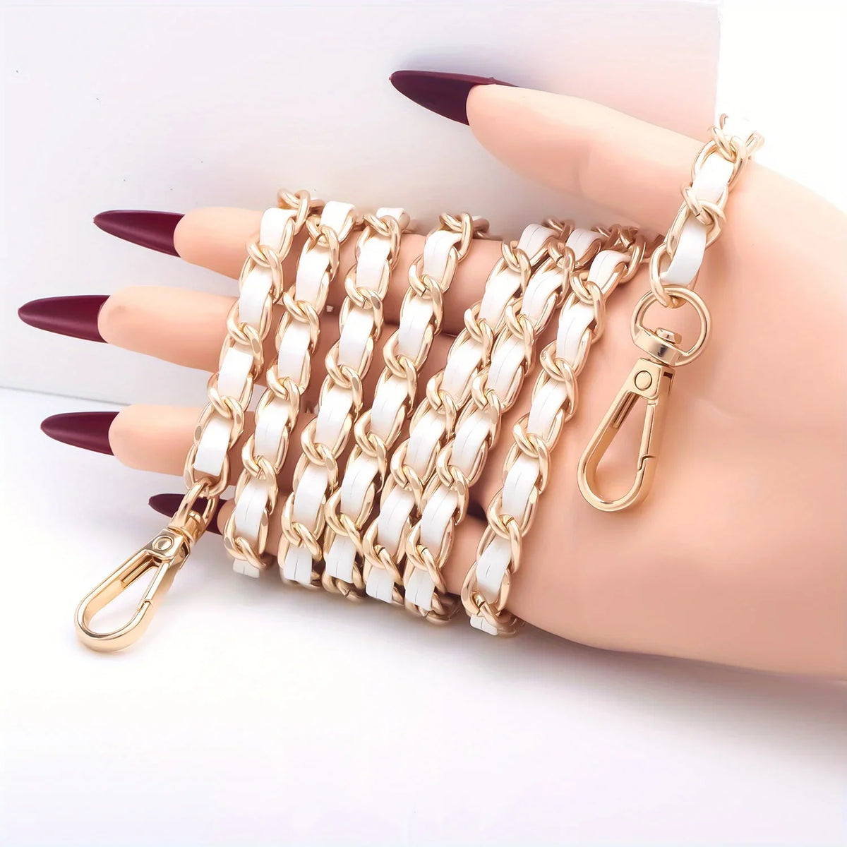 A hand displays a stack of gold-toned metal chain purse straps with white and gold links and a secure buckle.