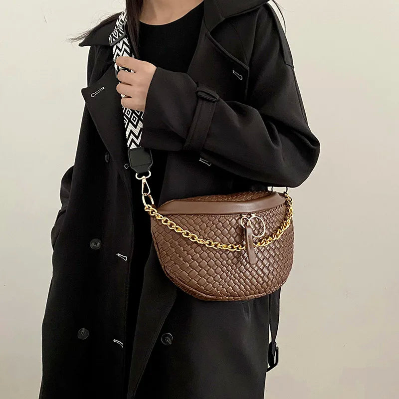 A woman wearing a black coat is holding a brown woven pattern waist bag with a gold chain strap.