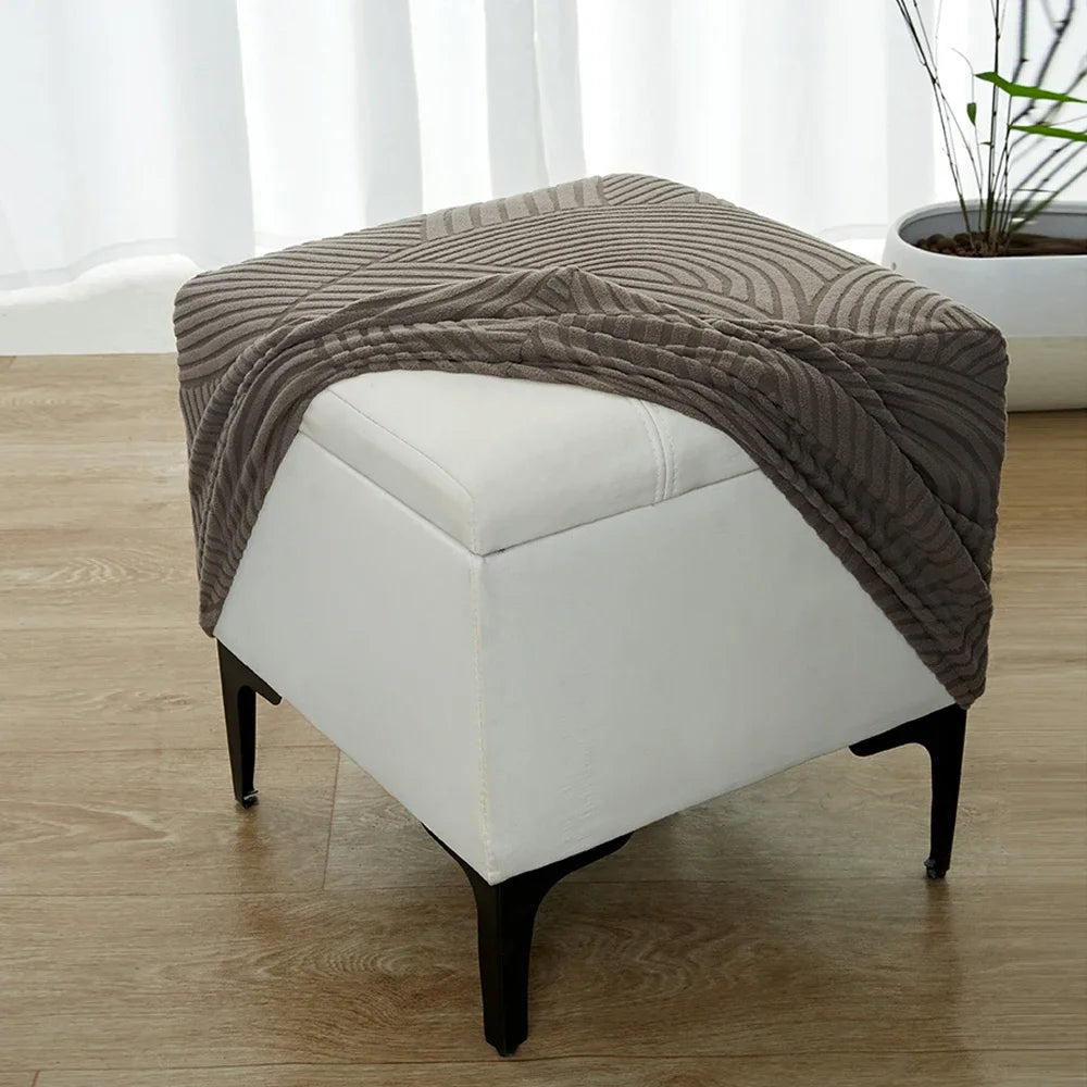 A beige geometric jacquard stretch ottoman cover draped over a white upholstered ottoman.