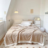 A beige fleece throw blanket is draped over a bed in a bright room.