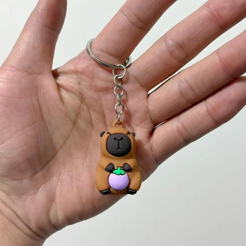 A hand holds an adorable capybara keychain charm with a purple fruit detail.