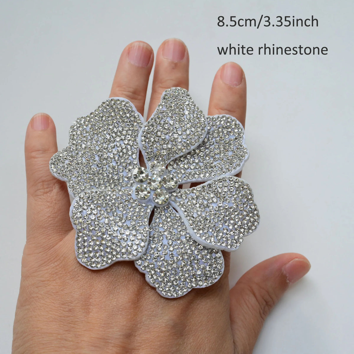 A hand holds a white rhinestone flower sew-on patch, measuring 8.5cm or 3.35 inches.