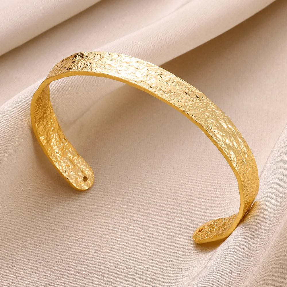 A close-up of an adjustable gold-tone stainless steel cuff bracelet lying on a beige fabric.